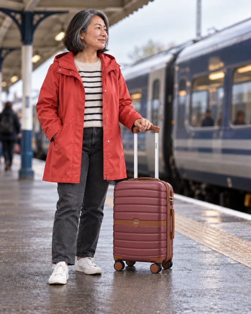 Woman in a coral jacket holding a compact burgundy and brown suitcase on a railway platform.