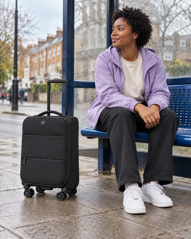 Woman in a lilac jacket seated beside a black soft-sided suitcase at a bus stop.